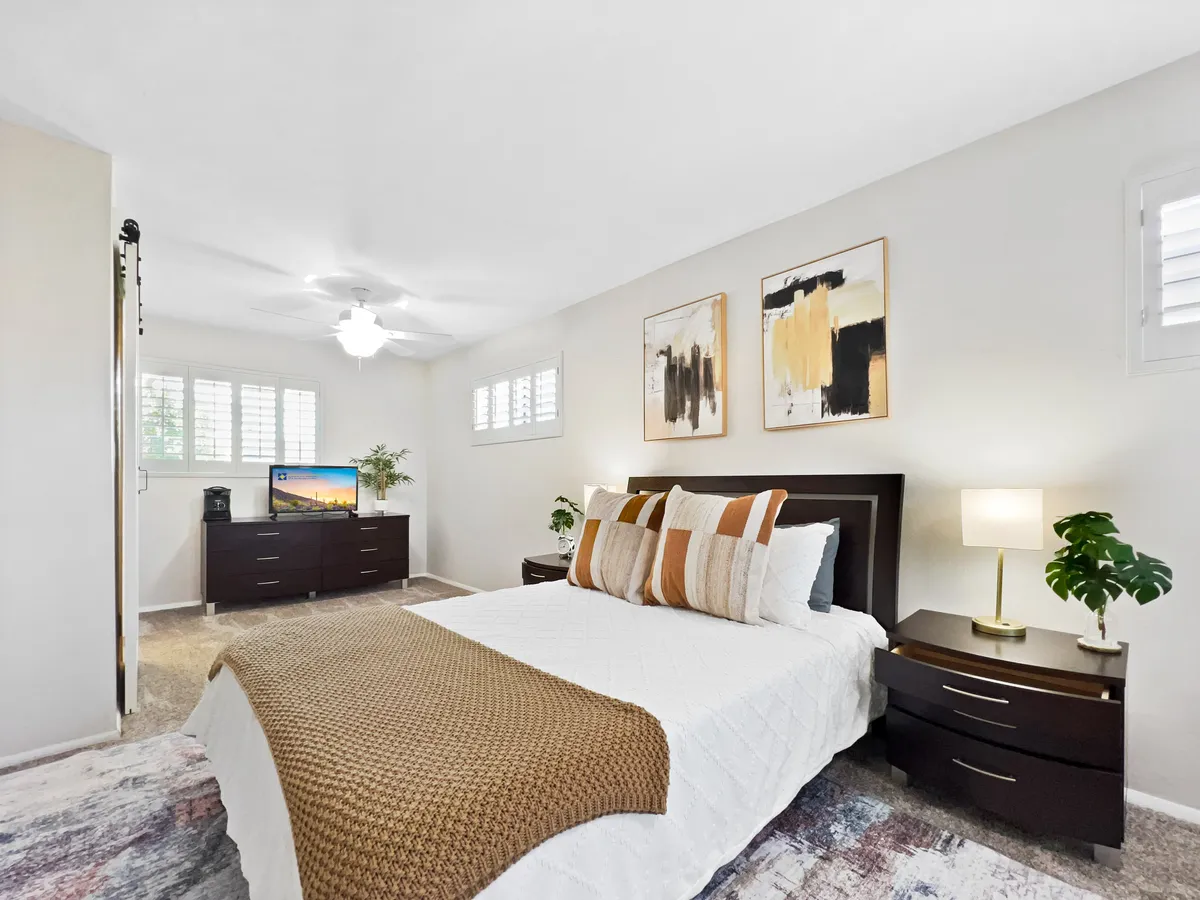The photo displays a calm and comfortable bedroom, including a bed with plush pillows, nightstands with ambient lighting, wall art, and a dresser with a streaming-ready TV and plant, embodying a cozy retreat.
