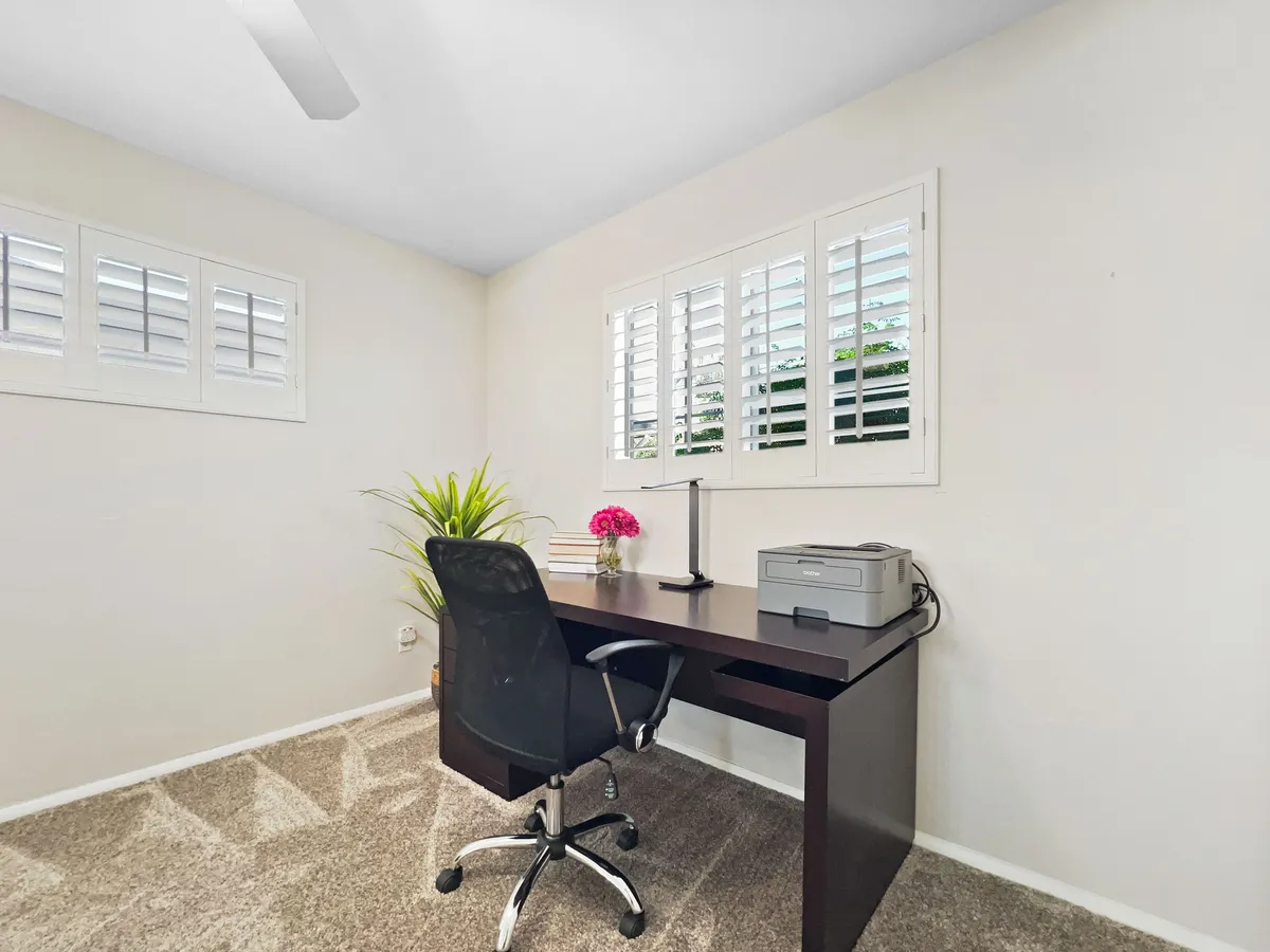 A focused workspace featuring a desk and ergonomic chair set by a window with shutters, complemented by natural light and a potted plant. A printer sits nearby for productivity, while the tranquil ambiance invites concentration.