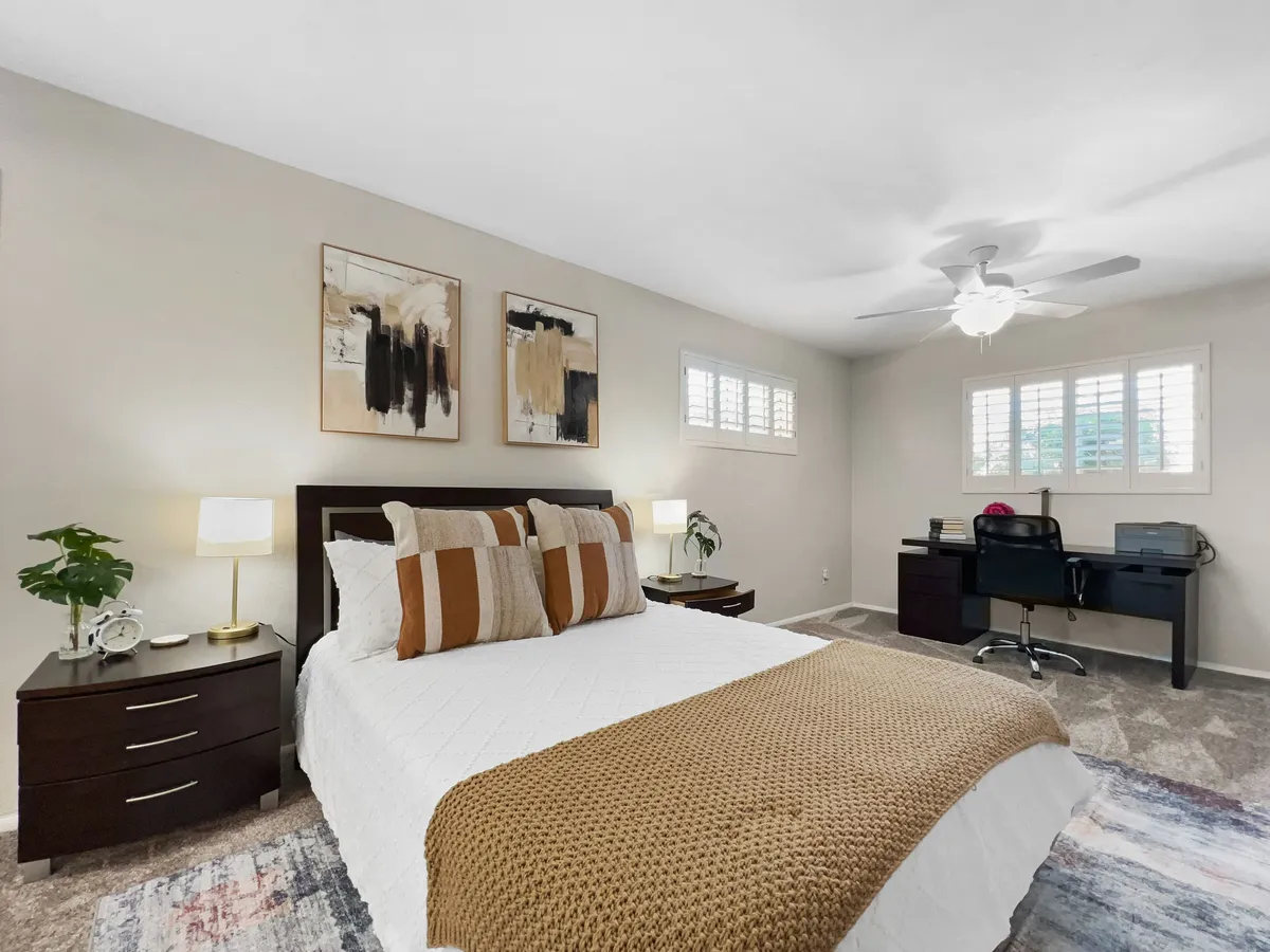 This image highlights a serene bedroom featuring a bed with decorative pillows, artwork on the wall, and a thoughtfully arranged desk area with a chair, creating a multi-functional space perfect for both relaxation and productivity.