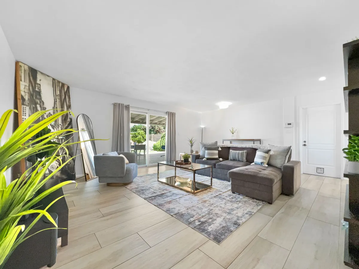 A comfortable sectional sofa set around a glass coffee table, with large windows providing natural light and views to the outside. The living room is accented by green plants and a decorative mirror, creating an inviting and relaxed atmosphere for gatherings or quiet mornings.