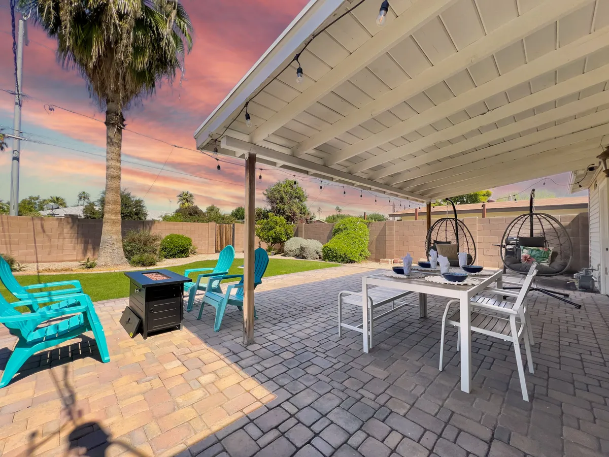 An inviting backyard view featuring hanging chairs, a dining table, a vibrant fire pit, and a backdrop of palm trees and colorful seating. The setup radiates a sense of social gathering, leisure, and connection to the outdoors.