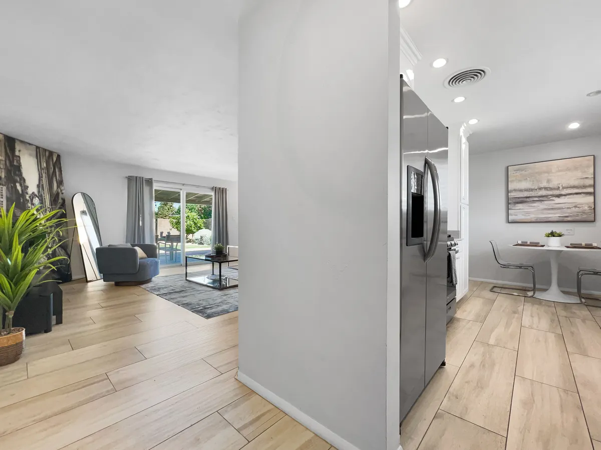 A wide-angle interior view that captures the seamless connection between the kitchen, dining area, and living room. The image highlights a modern refrigerator, inviting dining table with chairs, comfortable living room seating, mirrored wall decor, and a touch of greenery from an indoor plant. There's a sense of openness and flow that demonstrates the open-concept layout.